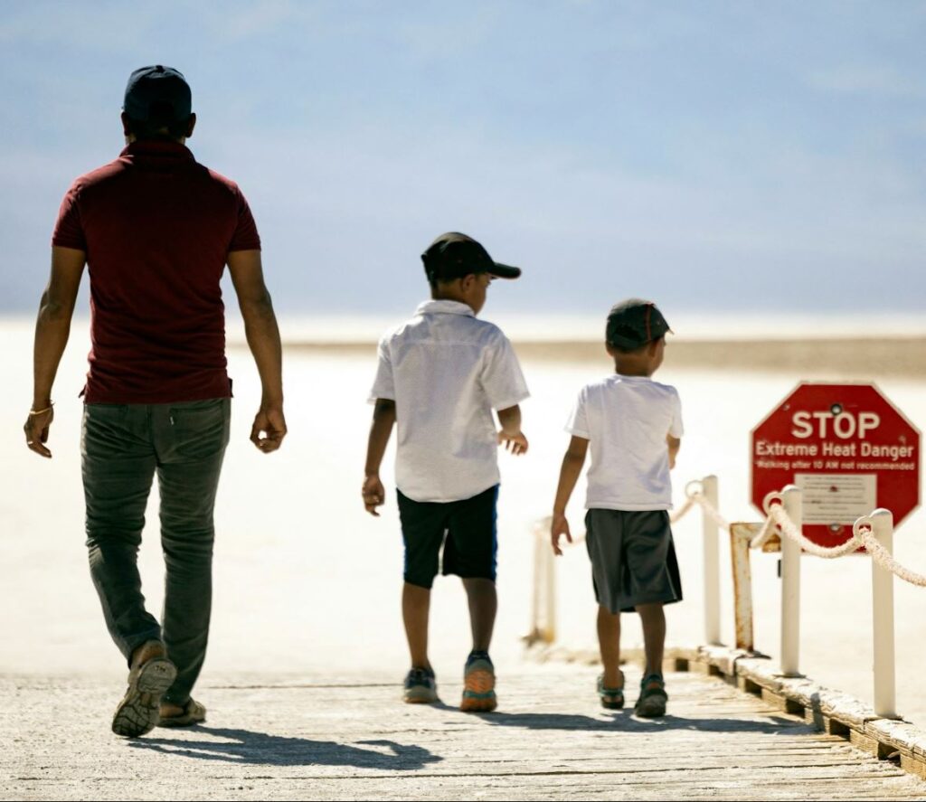 Photograph of a man and his son's entering the desert, next to a sign warning of dangerous heat